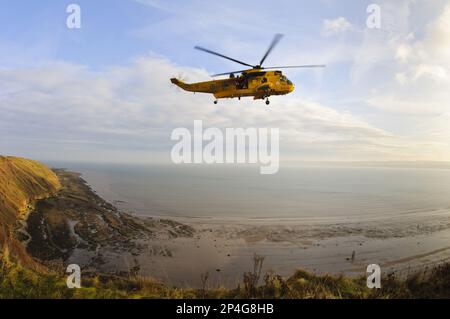 Hélicoptère de sauvetage RAF Westland Sea King avec membres d'équipage debout dans une porte ouverte en vol sur les côtes rocheuses et les falaises, vue depuis le sommet de la falaise à Banque D'Images