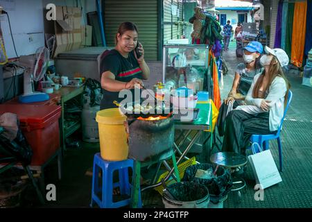 Un vendeur de nourriture de rue dans le marché central de Phnom Penh, au Cambodge, parle sur son téléphone mobile alors qu'elle frite des gâteaux sucrés et deux jeunes clients attendent. Banque D'Images