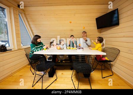 La famille avec quatre enfants mange des fruits dans une maison de campagne en bois le week-end. Le père et le fils jonglent avec les mandarines. Banque D'Images