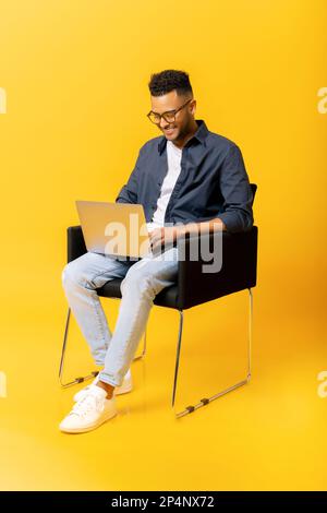Un Indien heureux regardant l'écran de l'ordinateur portable, en tapant du contenu d'information, en utilisant l'ordinateur pour travailler, assis au fauteuil isolé sur fond jaune. Recherche dans le concept Internet Banque D'Images