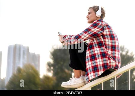 Femme assise sur un garde-corps en ville et écoutant de la musique à l'aide d'un casque. Banque D'Images