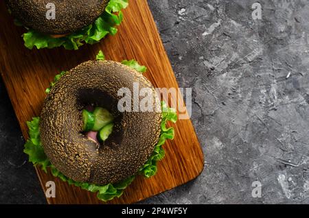 Sandwiches bagels noirs avec concombres, salade et jambon sur bois et fond sombre, délicieux en-cas ou petit déjeuner Banque D'Images