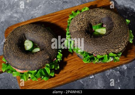 Sandwiches bagels noirs avec concombres, salade et jambon sur bois et fond sombre, délicieux en-cas ou petit déjeuner Banque D'Images