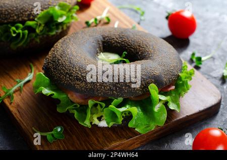 Sandwiches bagels noirs avec concombres, salade et jambon sur bois et fond sombre, délicieux en-cas ou petit déjeuner Banque D'Images