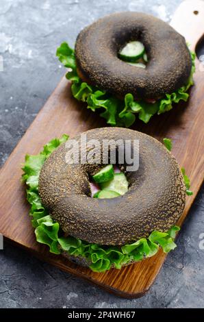 Sandwiches bagels noirs avec concombres, salade et jambon sur bois et fond sombre, délicieux en-cas ou petit déjeuner Banque D'Images