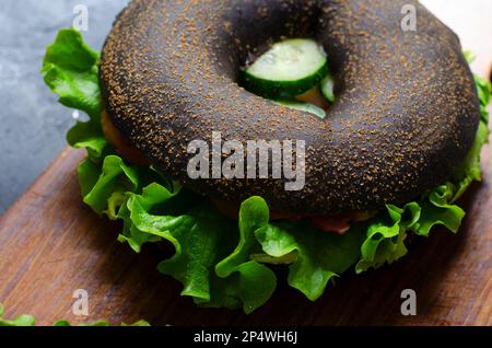 Sandwiches bagels noirs avec concombres, salade et jambon sur bois et fond sombre, délicieux en-cas ou petit déjeuner Banque D'Images