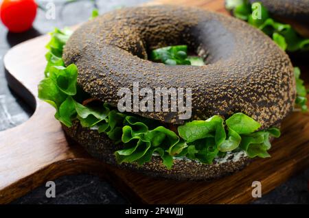 Sandwiches bagels noirs avec concombres, salade et jambon sur bois et fond sombre, délicieux en-cas ou petit déjeuner Banque D'Images