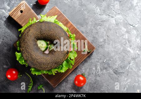 Sandwiches bagels noirs avec concombres, salade et jambon sur bois et fond sombre, délicieux en-cas ou petit déjeuner Banque D'Images