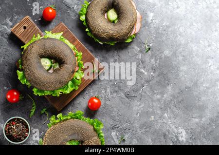 Sandwiches bagels noirs avec concombres, salade et jambon sur bois et fond sombre, délicieux en-cas ou petit déjeuner Banque D'Images