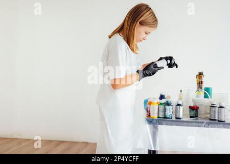 Une jeune fille adolescente dans un tablier blanc et des gants noirs tient un pot de peinture acrylique. Jeune artiste dans un atelier d'art. Il y a des peintures, des brosses et des tasses Banque D'Images
