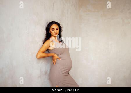 Un portrait de belle jeune femme enceinte. La femme Brunette en robe beige se tient contre un mur léger. Studio photo d'une femme qui attend un b Banque D'Images