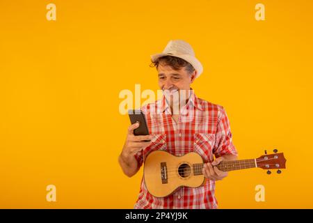 Homme habillé d'une manière gaie et jeune en utilisant son téléphone mobile et portant un ukulele Banque D'Images