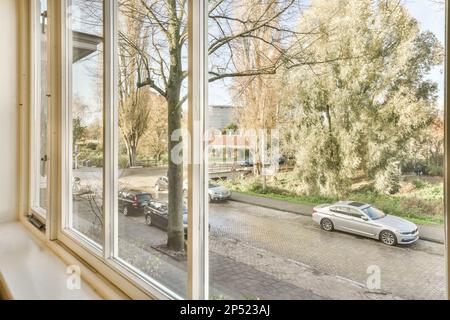 un espace extérieur avec des voitures garées dans la rue et des arbres de chaque côté de la route, vu par de grandes fenêtres Banque D'Images