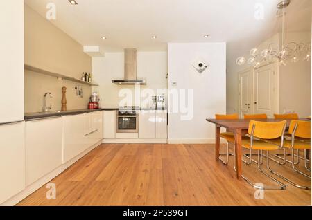 un coin cuisine et salle à manger dans un petit appartement avec parquet, armoires blanches, appareils en inox et chaises jaunes Banque D'Images