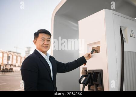 Portrait de l'homme beau en costume noir élégant en utilisant la carte de crédit tout en payant pour charger la voiture électrique à la station extérieure. Technologie moderne dans l'industrie automobile et achat sans contact. Banque D'Images