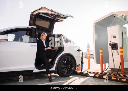 Une femme d'affaires caucasienne boit un café à emporter et travaille sur un ordinateur portable sans fil tout en chargeant la batterie de sa voiture électrique de luxe. Femme bien habillée qui fait des affaires à la station publique d'EV. Banque D'Images