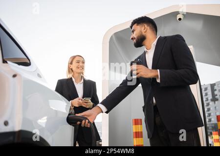 Femme caucasienne souriante et homme indien élégant vêtu d'un vêtement habillé avec un smartphone et buvant du café sur une station EV. Deux collègues professionnels discutant du travail tout en chargeant la voiture. Banque D'Images