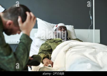 Un jeune soldat afro-américain blessé en uniforme militaire allongé dans le lit d'hôpital tandis que son ami inquiet était assis près de chez lui Banque D'Images