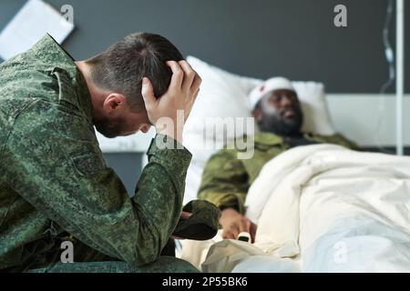Un jeune soldat inquiet en camouflage touche la tête tout en étant assis par son ami dans le lit d'hôpital après une blessure pendant des activités militaires Banque D'Images