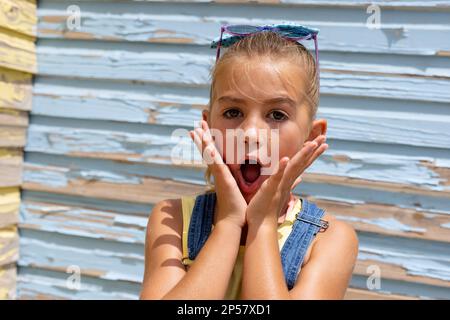 Portrait d'une jeune fille biraciale surprise tenant son visage avec la bouche ouverte sur le mur bleu Banque D'Images