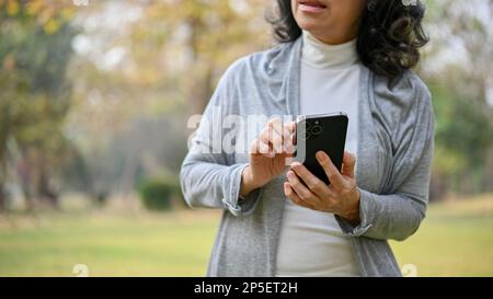 Image rognée d'une femme asiatique de 60 ans utilisant son téléphone, envoyant un message texte à quelqu'un en marchant dans le parc. Banque D'Images