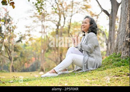 Vue latérale d'une femme asiatique heureuse et charmante de 60 ans vêtue de vêtements décontractés à l'aide de son téléphone tout en se relaxant et assis sur l'herbe sous un arbre dans un parc Banque D'Images