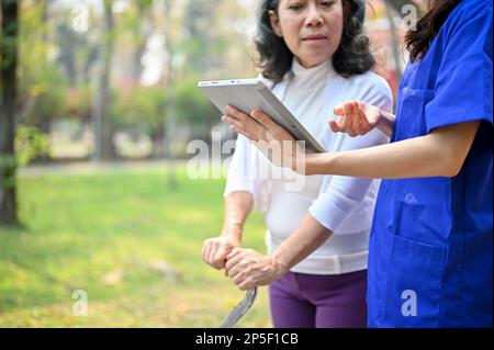 Image rognée d'une infirmière asiatique ou d'un soignant parle et montre quelque chose sur son écran de tablette à une vieille dame tout en marchant dans le parc tog Banque D'Images