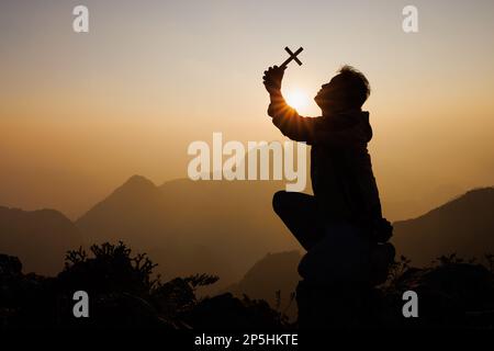 Silhouette d'un homme avec un crucifix dans ses mains priant à Dieu. Tandis que le soleil se lève, félicitez la religion. Foi suprême dans le Dieu du succès. ADV Banque D'Images