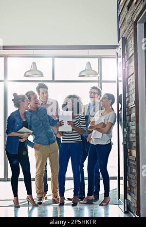 Nous travaillons dur, mais nous avons du plaisir au faire. un groupe de collègues en train de rire debout devant une fenêtre dans un bureau. Banque D'Images