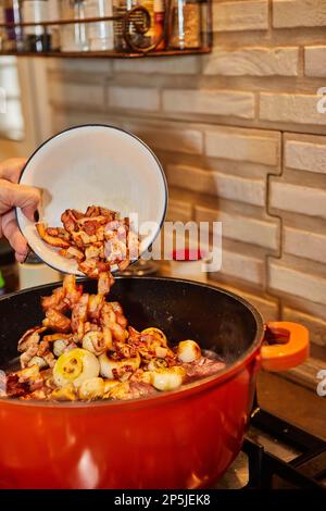 Le chef ajoute du bacon au poulet mariné dans du vin rouge avec du romarin dans une casserole sur une cuisinière à gaz Banque D'Images