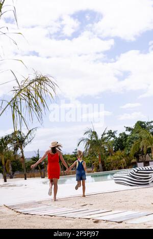 Bonne mère et fille biraciale marchant et tenant les mains près de la piscine Banque D'Images