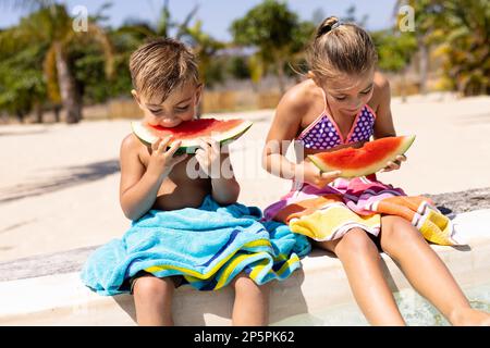 Joyeux frère biracial et sœur mangeant de la pastèque au bord de la piscine Banque D'Images