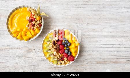 Bols à smoothie à la mangue saine de Ywo avec myrtilles, framboises, kiwi et granola. Scène au-dessus de la vue sur un arrière-plan en bois gris clair. Banque D'Images