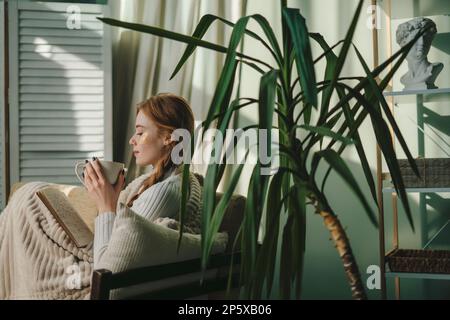 Sur les côtés, une jeune femme boit du café et lit un livre assis sur un canapé tout en restant à la maison, passant son temps libre dans le salon, à l'intérieur. Personnes Banque D'Images