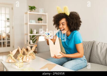 Femme noire préparant des cadeaux de Pâques à la maison, emballage de remplissage avec des œufs de chocolat Banque D'Images