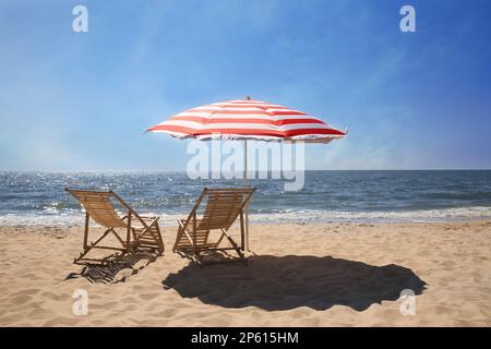 Chaises longues près d'un parasol rouge et blanc sur un bord de mer sablonneux Banque D'Images