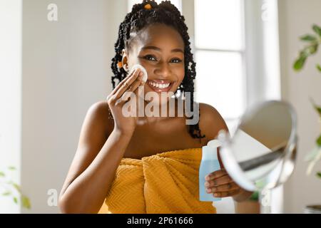 Belle femme noire tenant le flacon avec l'eau micellaire et tampon en coton pour enlever le maquillage, souriant à l'appareil photo Banque D'Images
