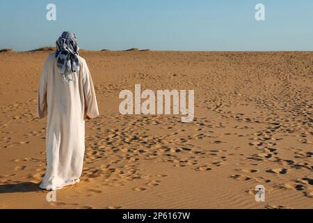 Homme en vêtements arabes marchant dans le désert le jour ensoleillé, vue arrière Banque D'Images