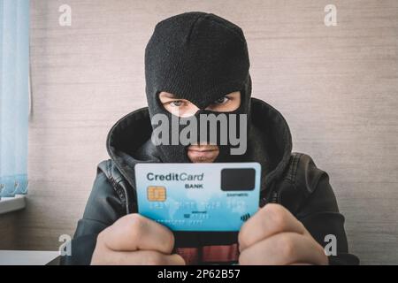 Criminel masqué qualifié utilisant une carte de crédit volée pour acheter des choses en ligne. Un voleur dans un masque noir tient une carte bancaire volée dans ses mains. Banque D'Images