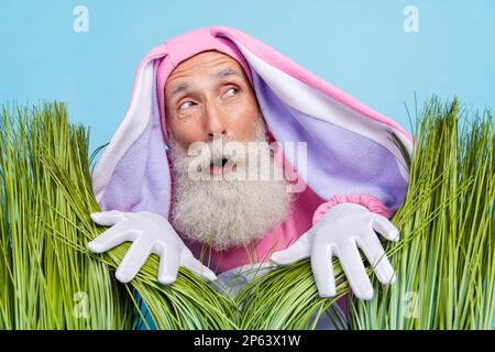 Photo de funky impressionné penseur gars habillé bunny kigurumi cacher l'herbe regardant vide espace isolé couleur bleu arrière-plan Banque D'Images