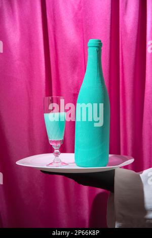 Garçon en gants noirs tient un plateau blanc avec un verre de vin turquoise et une bouteille de champagne. Arrière-plan des rideaux magenta. Banque D'Images