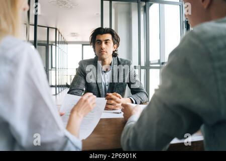 Portrait d'un homme confiant candidat à un emploi en tenue officielle interviewé par deux conseillers en RH méconnaissables lors d'une réunion au siège social. Banque D'Images