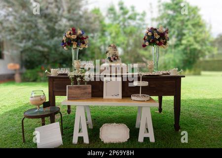 Gâteau de mariage sur une ancienne table en bois situé dans un jardin avec un fond d'arbres verts pendant une journée ensoleillée, décoré avec des bouquets de roses et des assiettes Banque D'Images