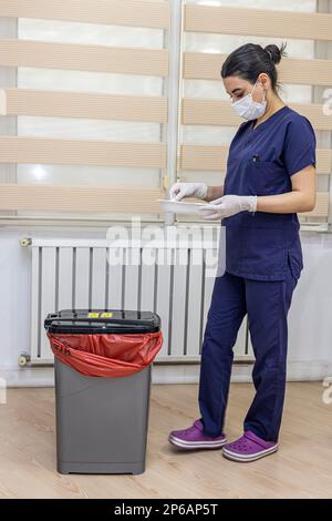 L'infirmière jette les déchets médicaux dans la poubelle de déchets médicaux du bureau. Une infirmière porte un sac poubelle rouge. Femme de ménage et corbeille des infections à l'hôpital. Infecti Banque D'Images