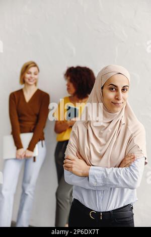 Belle femme d'affaires arabe en hijab regardant la caméra et souriant avec des collègues multiethniques en arrière-plan. Banque D'Images