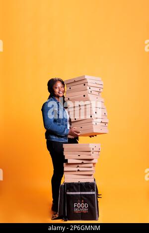 Livreur souriant tenant la pile pleine de boîtes de pizza se préparant à livrer aux clients pendant l'heure du déjeuner. Restaurant emloyé avec sac à dos thermique à emporter pour commander des plats à emporter Banque D'Images