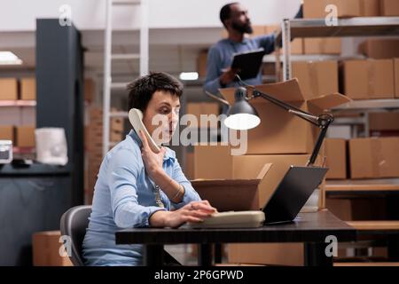 Un responsable caucasien parlant au téléphone fixe avec un superviseur à distance pour discuter d'un problème de logistique de transport tout en regardant le stock de fret sur ordinateur portable. Employé travaillant à la livraison de la commande dans l'entrepôt Banque D'Images