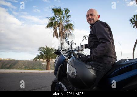 Motard assis sur son scooter avec un casque à la main dans un endroit tropical, photo avec espace copie. Banque D'Images