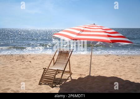 Chaise longue près d'un parasol rayé rouge et blanc sur un bord de mer sablonneux Banque D'Images
