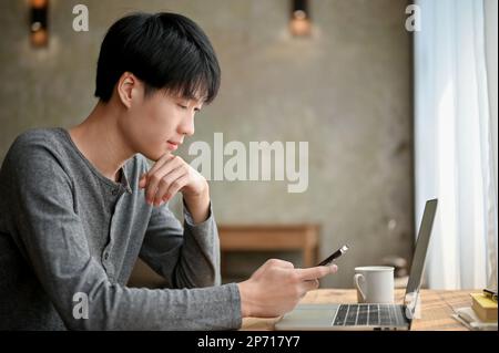 Image de côté d'un jeune asiatique travailleur indépendant ou étudiant pensivement en train de penser quelque chose en regardant l'écran de son smartphone, remo Banque D'Images